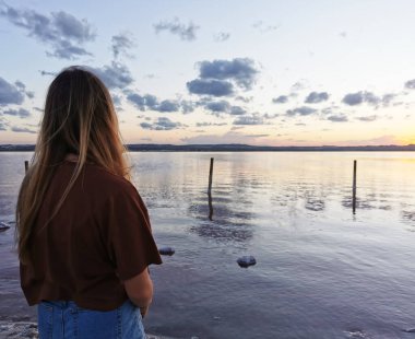Gölün kıyısında genç bir kız gün batımına ve göldeki bulutların yansımasına bakıyor.
