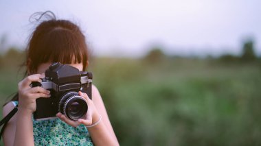 Küçük kız elinde orta formatlı film kamerası ve yeşil alan arka planlı günbatımı manzarasının fotoğrafını çekiyor.