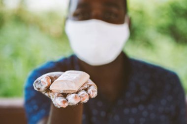 Afrikalı bir adam, yüz maskesi takmış ıslak bir sabun parçası tutuyor ve gösteriyor.