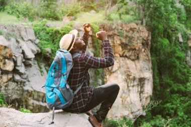 Afrikalı gezgin kayalıklarda oturuyor ve sırt çantasıyla kamerayı tutuyor.