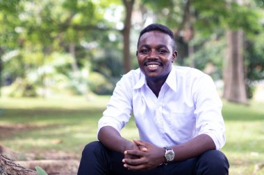 Afrikalı iş adamı oturmuş yeşil doğayla iç içe gülümsüyor.