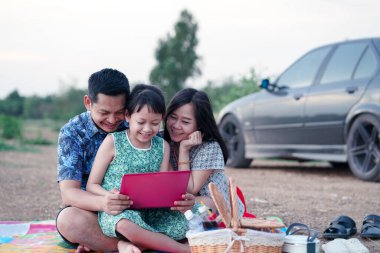 Asyalı bir aile piknik yapıyor. Parkta dizüstü bilgisayar izliyor. Gülümsüyor ve mutlu.