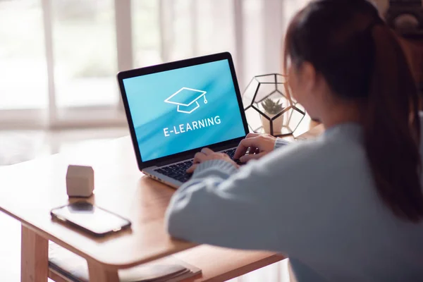 Online education, e-learning. Young woman is sitting at table, working on computer laptop with inscription on screen e-learning and image of square academic cap, distance training, copy space.
