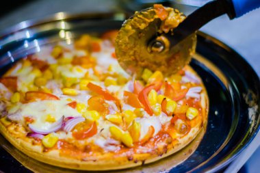 Cutting cheese pizza with Pizza cutter, with tomato, onion, corn toppings, with selective focus in lockdown.