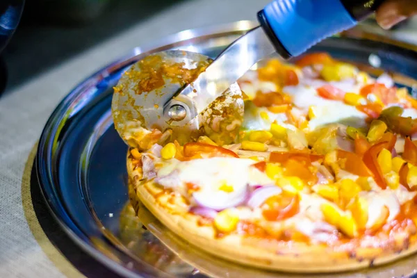 Cutting cheese pizza with Pizza cutter, with tomato, onion, corn toppings, with selective focus in lockdown.