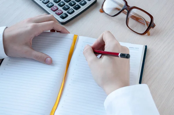 Beyaz gömlekli bir kız defterine kurşun kalemle, masaya hesap makinesi ve gözlük yazar.