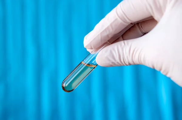 A hand in a disposable glove holds a glass tube with a transparent yellow liquid on a blue background