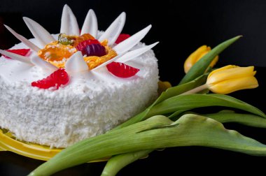 Beyaz pasta, taç yaprakları ve jöle ile süslenmiş, ve siyah arka planda sarı laleler.