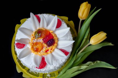 Beyaz pasta, taç yaprakları ve jöle ile süslenmiş, ve siyah arka planda sarı laleler.