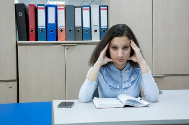 Ofis, esmer kızın başı ağrıyor, kız masada oturuyor ve elleriyle başını tutuyor.