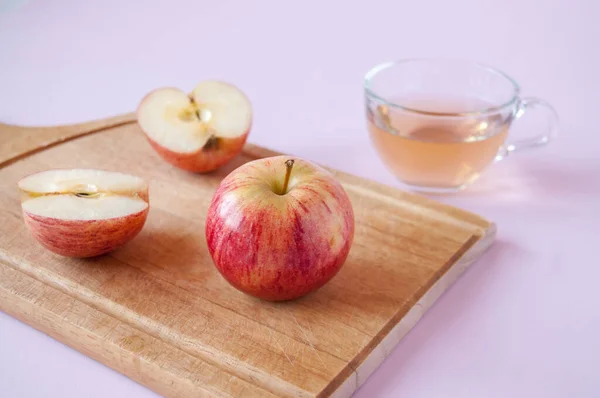 A whole and cut red Apple on a wooden Board and a mug of Apple juice on a pink background
