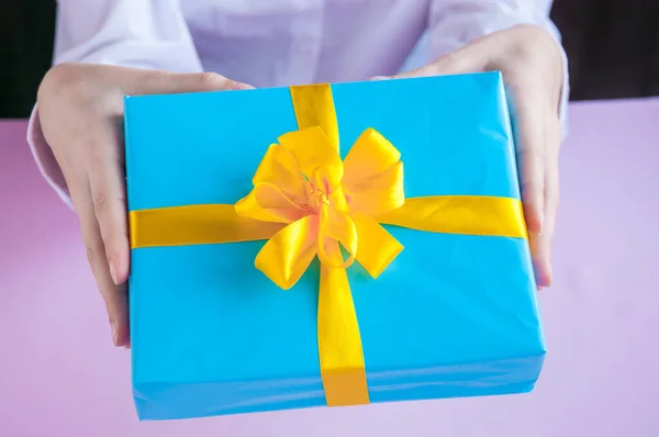 Female hands holding a gift in a blue box with a yellow ribbon on a pink background