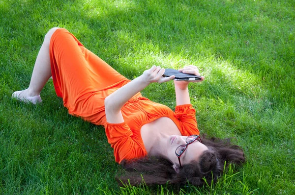 Turuncu elbiseli esmer, akıllı telefonlu çimenlerin üzerinde yatıyor.