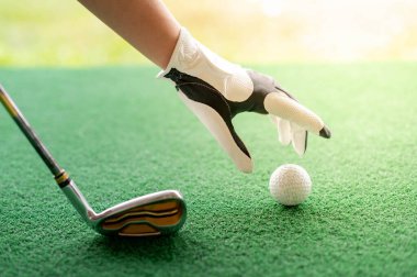Sol eliyle beyaz profesyonel eldivenler giyen kadın golf topunu ve golf sopasını çimlerin üzerine koyup golf topuna vurmaya hazırlanıyor..