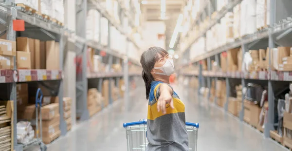 The asian girl wearing surgical mask pushing the trolley cart shopping the decorate funiture for interior inside the house in the big warehouse store.