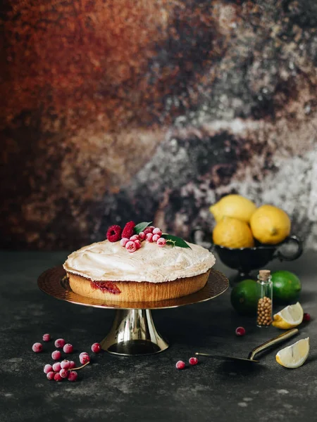 Lemon pie with berries. On a golden box for cakes. Nearby are lemons and limes. Berries of raspberries and red currants. Background gray and brown. Home baking. Meringue. Ready at home.