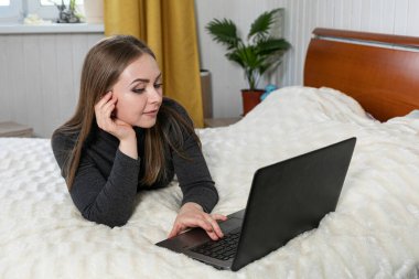 Kız dizüstü bilgisayarla yatakta yatıyor. Kızın beyaz teni ve sarı saçları var. Gri ceket ve kot pantolon. Beyaz oda. Tatil evi. Online eğitim. Mesafe öğrenme. Evde oturuyoruz..