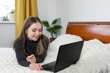 Kız dizüstü bilgisayarla yatakta yatıyor. Kızın beyaz teni ve sarı saçları var. Gri ceket ve kot pantolon. Beyaz oda. Tatil evi. Online eğitim. Mesafe öğrenme. Evde oturuyoruz..