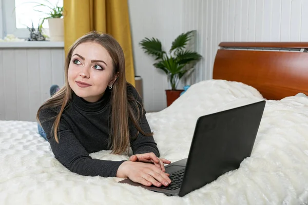 Kız dizüstü bilgisayarla yatakta yatıyor. Kızın beyaz teni ve sarı saçları var. Gri ceket ve kot pantolon. Beyaz oda. Tatil evi. Online eğitim. Mesafe öğrenme. Evde oturuyoruz..