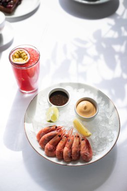 Restoranda taze deniz ürünleri. Beyaz tabakta buzlu karides. Beyaz masa örtüsü. Güneş parlıyor, bitkilerin gölgesi. Yakınlarda taze tutku meyveleriyle süslenmiş kırmızı bir kokteyl var. Bir restoran..