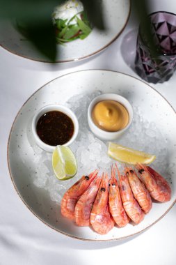 Fresh seafood in a restaurant. Shrimps on a white plate with ice. White tablecloth. Sauces, llama and lemon pussies. Nearby is a glass cup. A plate with salad is visible.