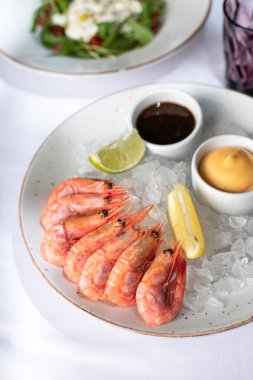 Fresh seafood in a restaurant. Shrimps on a white plate with ice. White tablecloth. Sauces, llama and lemon pussies. Nearby is a glass cup. A plate with salad is visible.