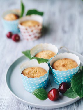 Ev yapımı pişirme. Mavi ambalajlı vişneli kekler tabakta. Arka planda yeşiller ve bulanık kekler var. Tatlılar taze böğürtlen ve yapraklarla süslenir. Ahşap arka plan. Yemek stili.