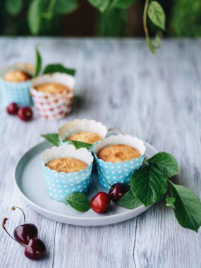 Ev yapımı pişirme. Mavi ambalajlı vişneli kekler tabakta. Arka planda yeşiller ve bulanık kekler var. Tatlılar taze böğürtlen ve yapraklarla süslenir. Ahşap arka plan. Yemek stili.