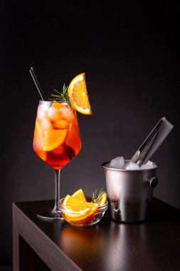 Aperol sprey kokteyli bir şarap bardağında servis edilir. Portakal ve biberiye dilimleri ile süslenir ve bir bar tezgahına yerleştirilir.