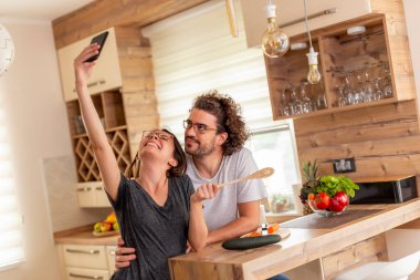 Çift mutfakta birlikte yemek hazırlarken, akıllı telefon kullanarak selfie çekerken eğleniyor.