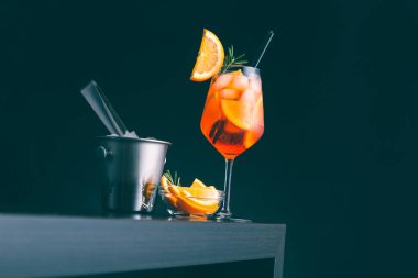 Aperol spreyli kokteyl, bir bar tezgahına yerleştirilmiş, bir dilim portakal ve biberiye ile süslenmiş, bol buzlu bir şarap kadehinde servis edilir.