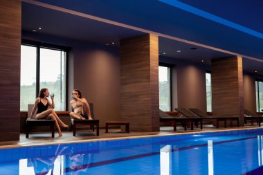 Güzel, genç ve aşık bir çift spa merkezinde dinleniyor, yüzme havuzunun kenarında güneşleniyorlar ve su içiyorlar, tatillerinin tadını çıkarıyorlar.