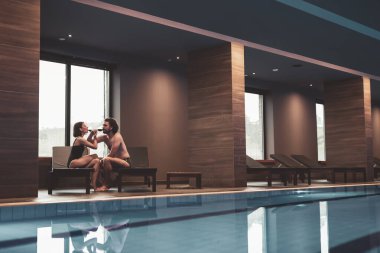 Aşık bir çift tatilin tadını çıkarıyor, spa merkezinin havuzunda dinleniyor, güneşli yataklarda oturuyor, sohbet ediyor, gülüyor ve şarap içiyor.