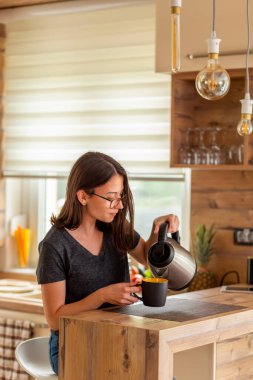 Güzel genç bir kadın mutfak tezgahında oturmuş, kendisine bir fincan sıcak kahve dolduruyor, sabahları evde boş vakitlerin tadını çıkarıyor.