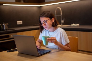 Pijama giyen, evde mutfak masasında oturan, geceleri geç saatlere kadar çalışan, dizüstü bilgisayar kullanan ve kahve içen serbest çalışan bir kadın.