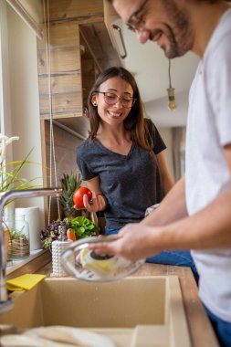 Genç aşıklar öğle yemeğinden sonra bulaşıkları beraber yıkıyorlar. Kadın tezgahta oturmuş gülümserken erkek bulaşıkları yıkıyor.