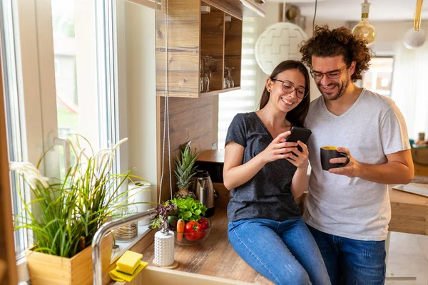美しい若いカップルは 朝のレジャー時間を楽しんで キッチンでコーヒーを飲み スマートフォンを使用してインターネットをサーフィンするのが大好きです — ストック写真