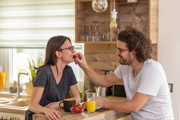 Young couple in love sitting at kitchen counter, drinking coffee and eating strawberries for breakfast