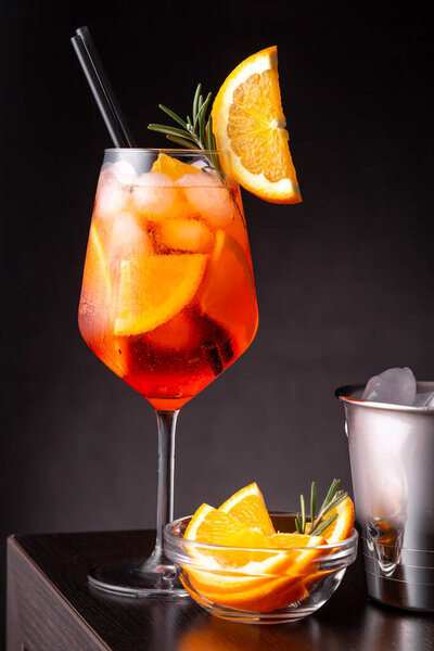 Aperol spritz cocktail served in a wineglass with lots of ice, decorated with slice of orange and rosemary branch, placed on a bar counter