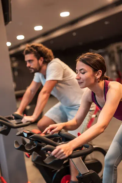 Handsome young people working out in gym, cycling on bike machines, doing cardiovascular training session