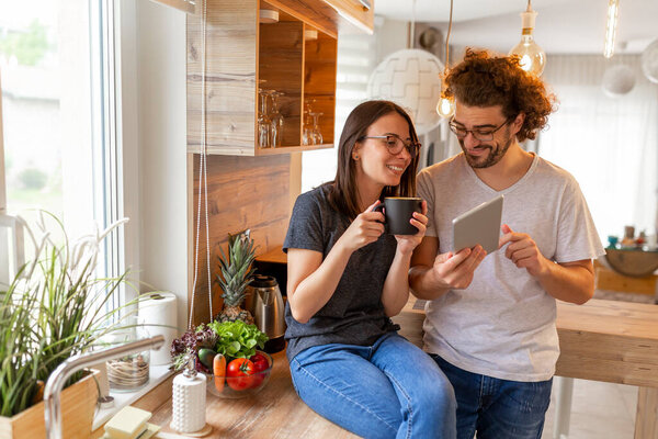 Couple in love drinking coffee and using a tablet computer in the kitchen, enjoying their leisure time together and relaxing at home