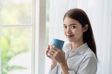 Mutlu Asyalı kadın sabah uyandıktan sonra bir fincan kahve içip camdan dışarı bakıyor.