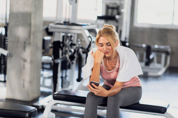 Portrait of fitness woman holding bottle of drinking water and using mobile phone in the gym