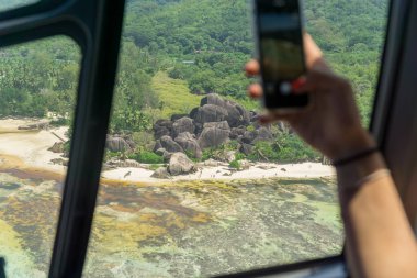 Aerial view in helicopter cabin of the tropical la digue Island. hand with a smartphone taking photo clipart