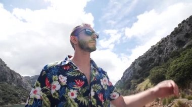 Havai gömlekli adam modelliği ve güneşli güzel bir günde güneş gözlüğünü çıkarması arkasında dağ manzarası olan Gorg Blau gözcüsü, Mallorca Adası, İspanya. Yüksek açı, seyahat + parallax hareketleri, yavaş çekim, HD.