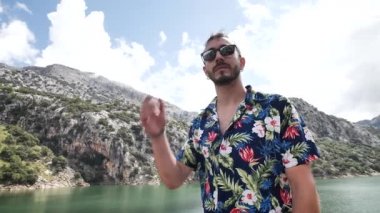 Havai gömlekli bir adam, güneş gözlüğünü çıkarıyor ve güzel güneşli bir günde kameraya gülümsüyor arkasında dağ manzarası olan Gorg Blau gözcü, Mallorca Adası, İspanya. Yüksek açı, seyahat + parallax hareketleri, yavaş çekim, HD.