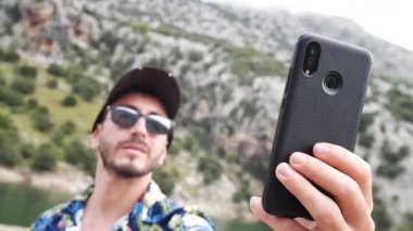 Hawaii gömlekli bir adam, güzel güneşli bir günde arkada dağ manzaralı bir selfie çekiyor, Gorg Blau gözcülük yapıyor, Mallorca adası, İspanya. Orta açı, seyahat + parallax hareketleri, yavaş çekim, HD.
