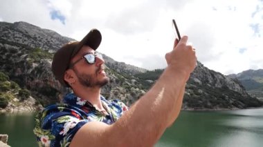 Hawaii gömlekli bir adam, güzel güneşli bir günde arkada dağ manzaralı bir selfie çekiyor, Gorg Blau gözcülük yapıyor, Mallorca adası, İspanya. Orta açı, seyahat + parallax hareketleri, yavaş çekim, HD.
