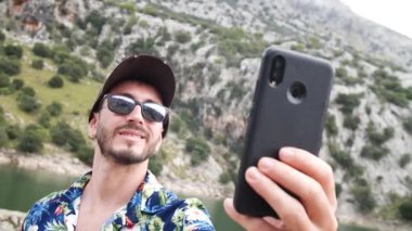 Hawaii gömlekli bir adam, güzel güneşli bir günde arkada dağ manzaralı bir selfie çekiyor, Gorg Blau gözcülük yapıyor, Mallorca adası, İspanya. Orta açı, seyahat + parallax hareketleri, yavaş çekim, HD.