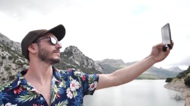 Hawaii gömlekli bir adam, güzel güneşli bir günde arkada dağ manzaralı bir selfie çekiyor, Gorg Blau gözcülük yapıyor, Mallorca adası, İspanya. Orta açı, seyahat + parallax hareketleri, yavaş çekim, HD.